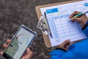a hand holds a clipboard with a Air Quality measuring table on it and is writing down measurements. Another hand holds a smartphone with and Air Quality measuring App on it. They are outside
